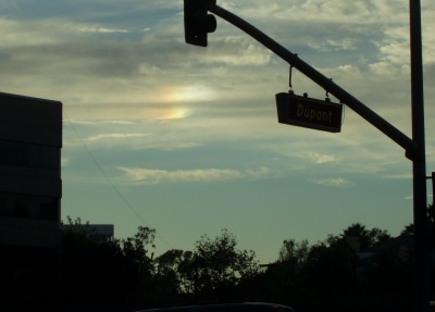 The long boom of a traffic signal and street sign are silhouetted against a blue-gray sky banded with clouds. There is a bright spot in the clouds, white fading to yellow and red toward the left.