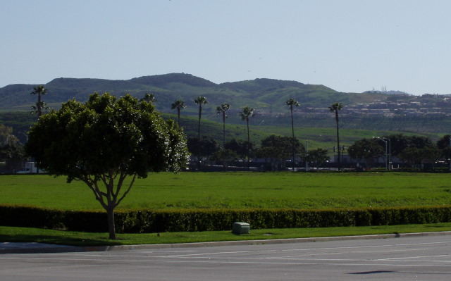 Street corner, facing a vacant lot covered with green grass, in front of green hills with a clutch of houses to the right.
