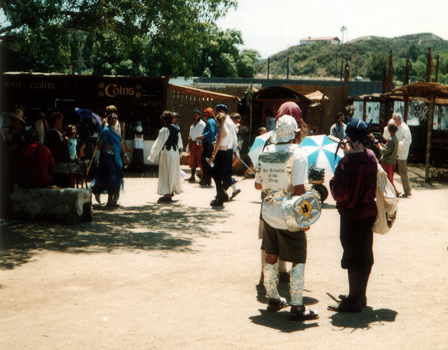 A man dressed in aluminum foil with a sign on his back reading Sir Reynolds of the Wrap, talking to other people dressed in more serious Renaissasnce Faire garb. More people are milling about in front of some shop stalls.