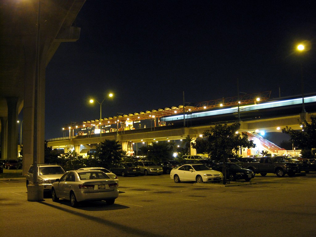 Elevated train station above a parking lot at night. A long streak of light indicates the windows of a train in motion.