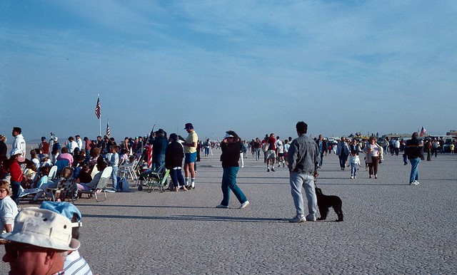 Crowd of people with lawn chairs facing the same way and walking aroundor walking around on the dirt.