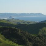 Green hills roll back to a half-moon shaped bay in the distance, an immense round rock near the shoreline. The rock is taller than the trio of smokestacks nearby. More hills are visible on the far side of the bay, and the hazy gray-white-blue sky above the brighter blue of the ocean.