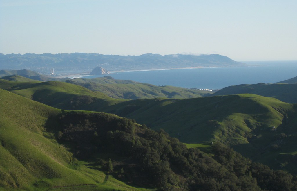 Green hills roll back to a half-moon shaped bay in the distance, an immense round rock near the shoreline. The rock is taller than the trio of smokestacks nearby. More hills are visible on the far side of the bay, and the hazy gray-white-blue sky above the brighter blue of the ocean.