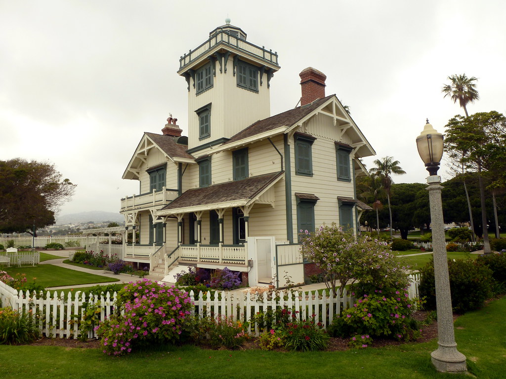 It's a cloudy day. Behind a white picket fence and green lawn, there's a whitewashed, gabled housel Most of it is two stories, but a square tower in the middle rises two more levels, and the tower is topped with a railing above which which you can just glimpse the top of a glass structure.
