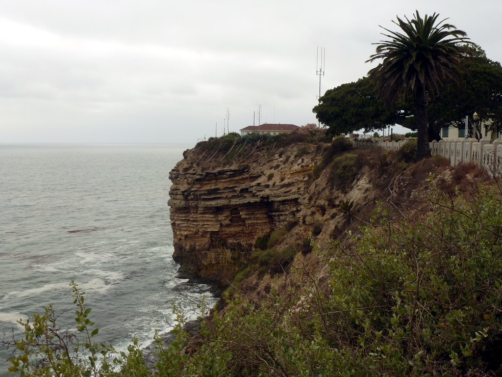 It's gloomy. The sky and sea are almost the same shade of gray. Looking along a steep cliffside toward a rock outcropping. It's high, maybe ten times as high as the low building perched on top of it, and grooves show dozens of layers stacked from the sea below to a bit below the top.