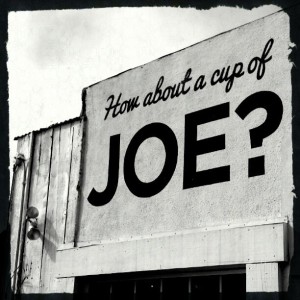 Stark black and white image of a sign painted directly on an outer wall. Wooden boards are visible next to it, and the top of a window frame below it. The sign reads "Hoa about a cup of" in cursive, and then in MUCH LARGER SANS-SERIF BLOCK LETTERS, "JOE."