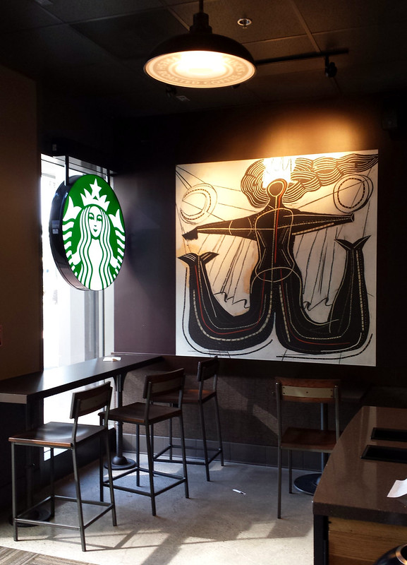 Empty corner of a coffee shop. To the left is a lit sign with Starbucks logo. Above is a circular ceiling lamp. On the wall is a stylized painting showing a mermaid with two tails and a circle on each side.