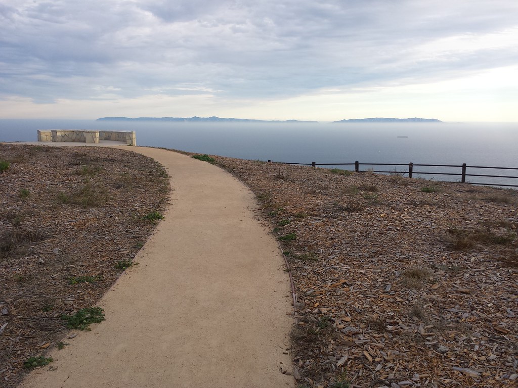 Dirt path curving away toward a hilltop with a bench. Only ocean and sky are visible beyond the hilltop