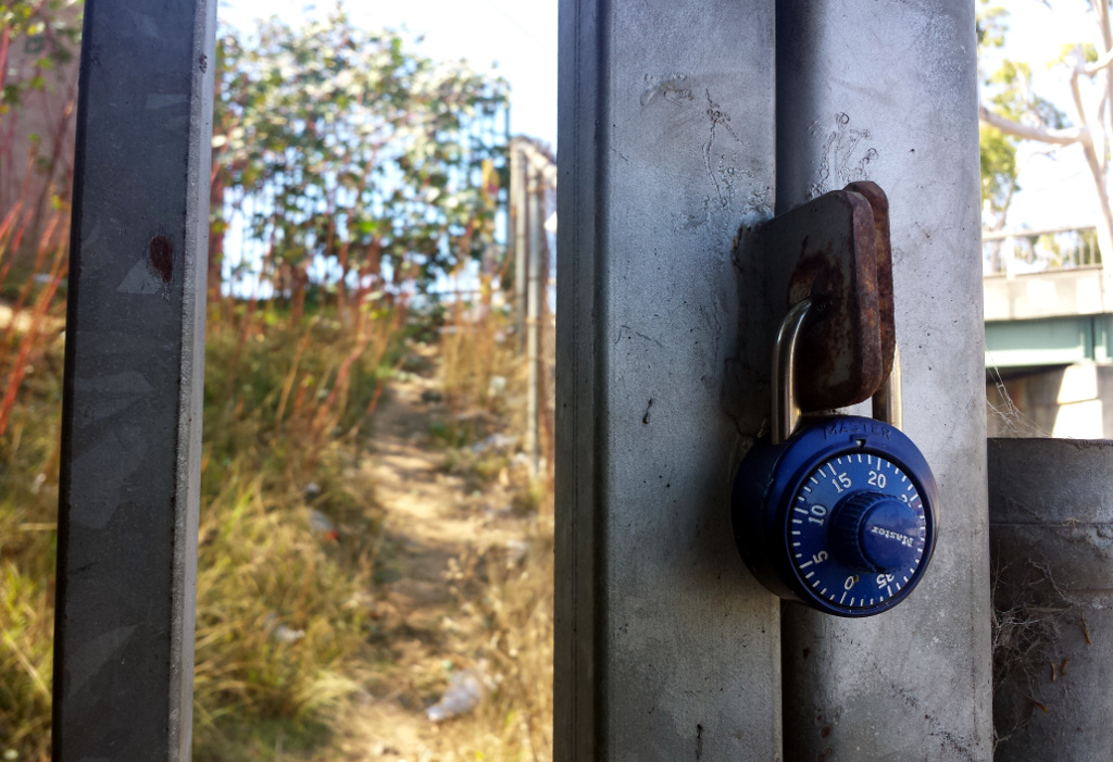 Close-up: looking between bars of a metal fence. 2 bars are locked together with a combination lock. Behind the gate we can see a blurry path uphill and scraggly bushes, with another fence at the top of the path.