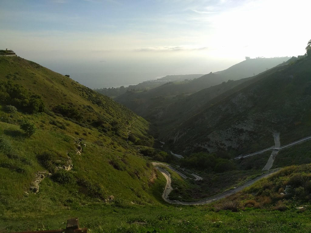 Looking down a green valley toward a hazy coastline. A path winds along the bottom, disappearing behind shadowed hills long before it reaches the sea.