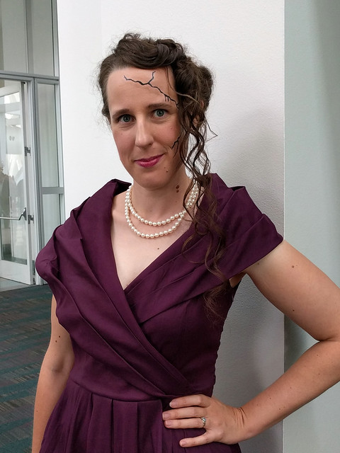 Whitney Frost Cosplay: Woman in a 1940s style purple dress, spidery cracks running down the side of her face, and hair with half a victory roll and curls on one side.