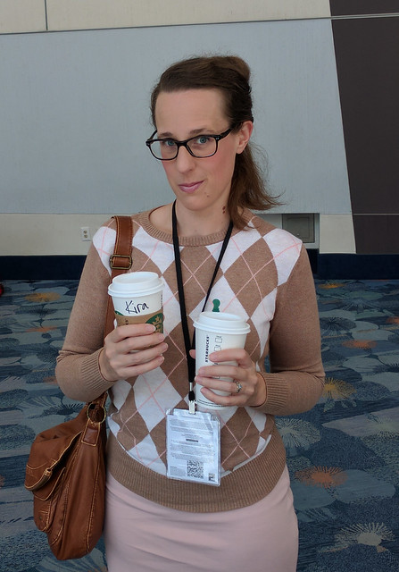 Kara Danvers cosplay: Woman wearing a harlequin sweater and glasses, carrying cardboard coffee cups, one labeled Kira.