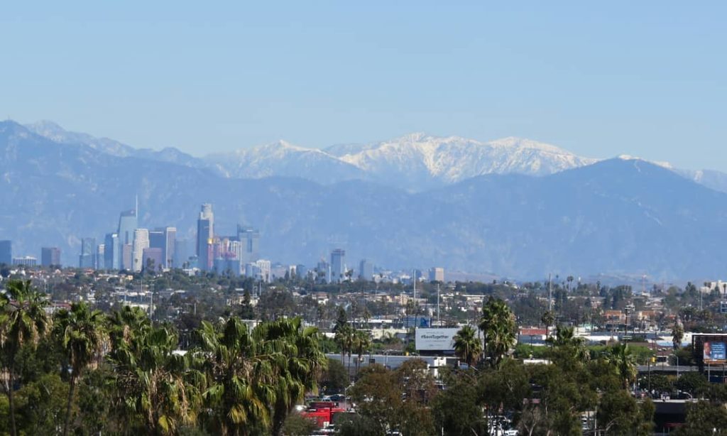 Hazy view of mountains with some snow on top, not much lower down. A city skyline is visible between the mountains and a canopy of trees and low buildings.