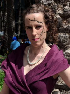 Woman in a purple dress, off-center 1940s hair, and a crack of darkness spidering its way down her face