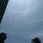 Thin clouds and contrails cross the sky. A bright rainbow-like ring circles a spot just out of view above the frame. A fainter rainbow-like line runs across the sky below it.