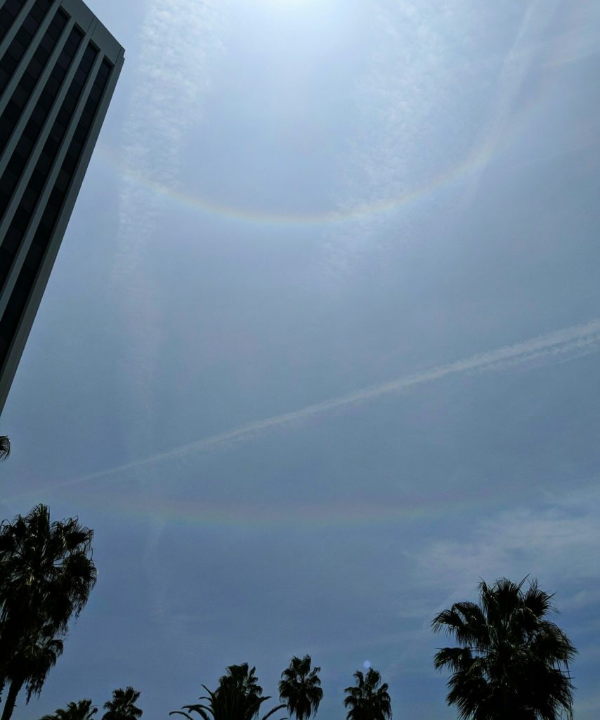 Thin clouds and contrails cross the sky. A bright rainbow-like ring circles a spot just out of view above the frame. A fainter rainbow-like line runs across the sky below it.