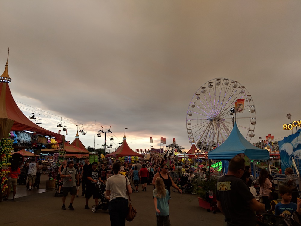 [Crowds at the fair, and rides, with smoke above and some blue sky in the distance.]