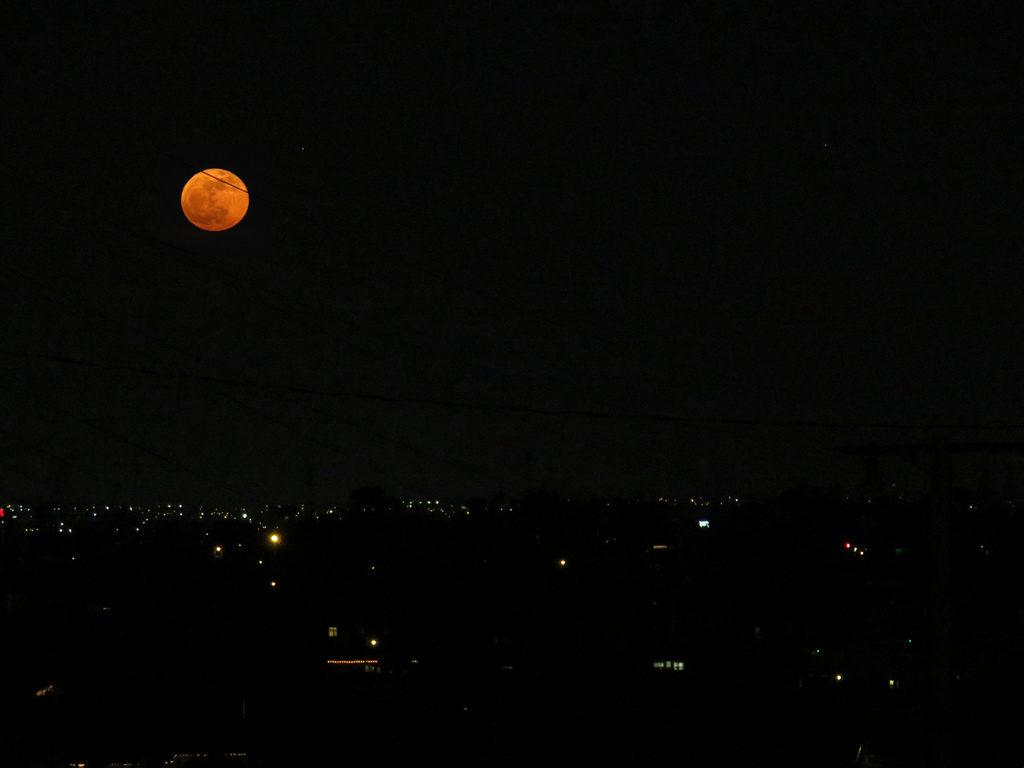 Orange moon in a night sky above a darkened horizon with city lights.