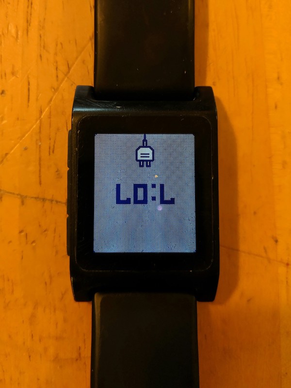 Pebble watch showing 7:07 and a charging icon. Upside-down so it looks like LOL.