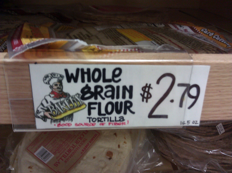 Grocery store shelf with a sign that says Whole Grain Flour Tortillas...but the G really looks like an 8 or a B.