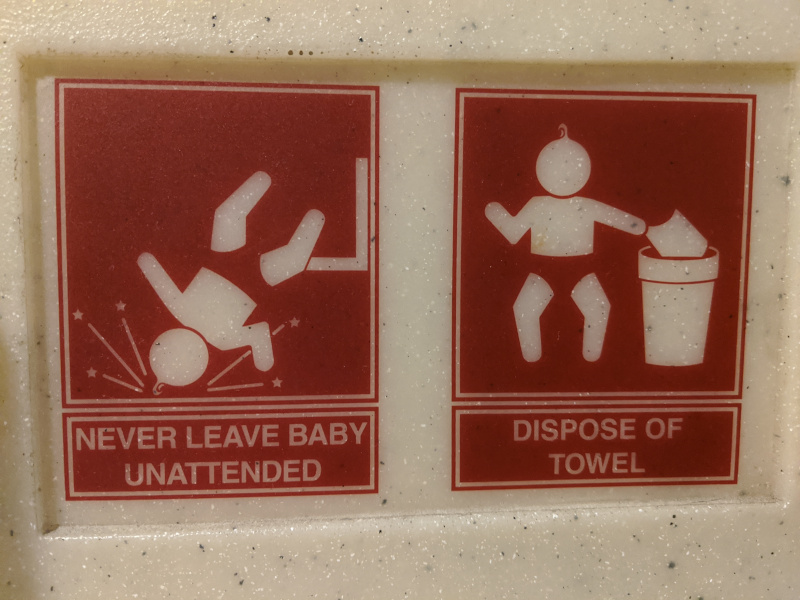 Signs on a changing table: Never Leave Baby Unattended (stick figure of a baby falling on its head). Dispose of Towel (stick figure of a baby carefully placing a towel in a trash can.