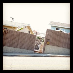 Fences, houses, and a gate on the side of a hill, tilted so that the sidewalk and street are horizontal.