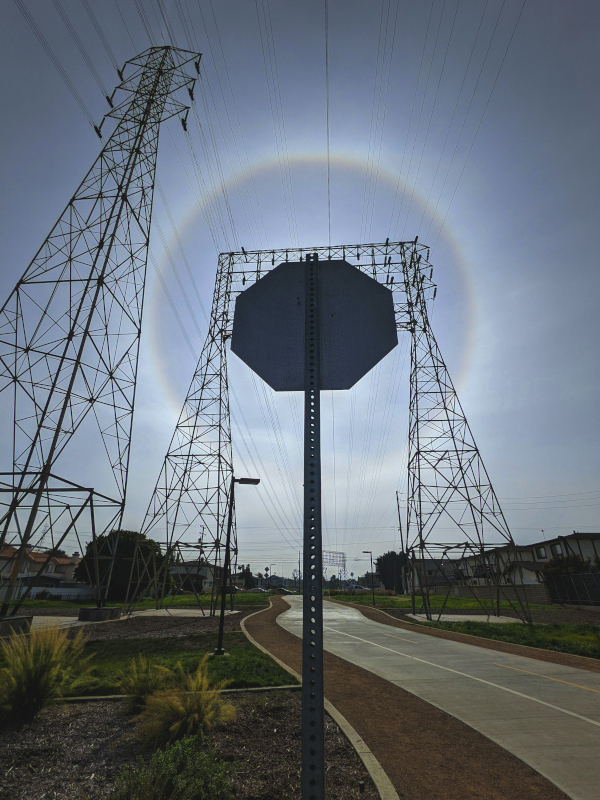 A bright circle in the sky surrounds the silhouette of a stop sign blocking the sun. Below it, a bike path winds into the distance and electrical towers stretch upward.