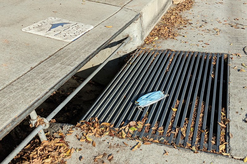 A discarded disposable face mask on top of a storm drain at the edge of a street.