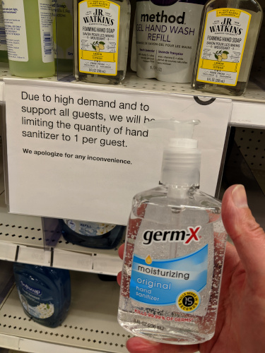 Holding a bottle of hand sanitizer in front of a store shelf with a sign saying that customers are limited to one bottle each due to high demand.