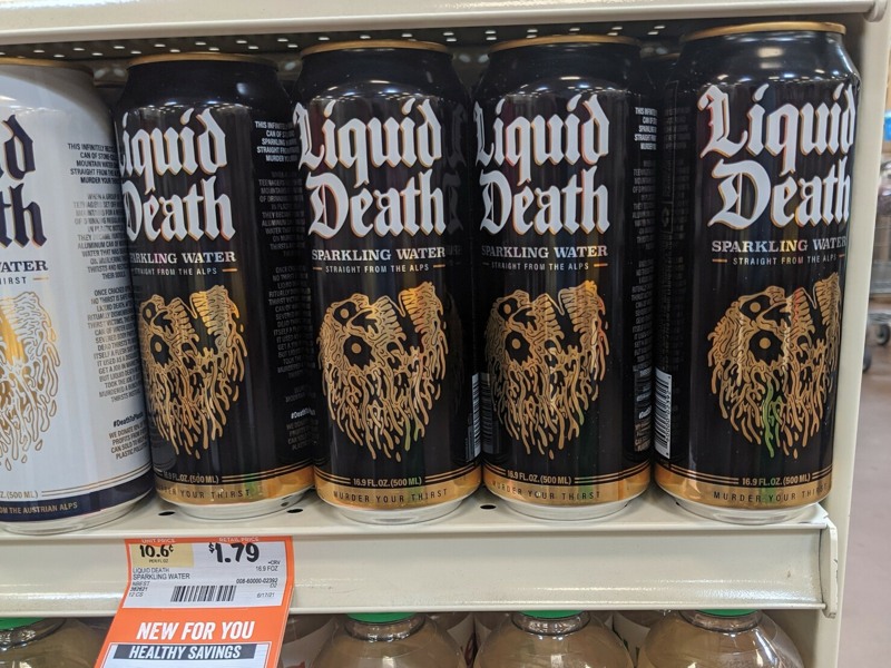 "Drink cans on a shelf labeled Liquid Death in calligraphy with a flaming skull. The shelf has a price label with a discount labeled Healthy Savings.