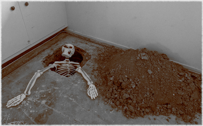 Sepia-stylized photo of a plastic skeleton climbing out of a 1.5x1.5 foot hole in a concrete floor between a set of clearly-indoor cabinets and a big pile of dirt.