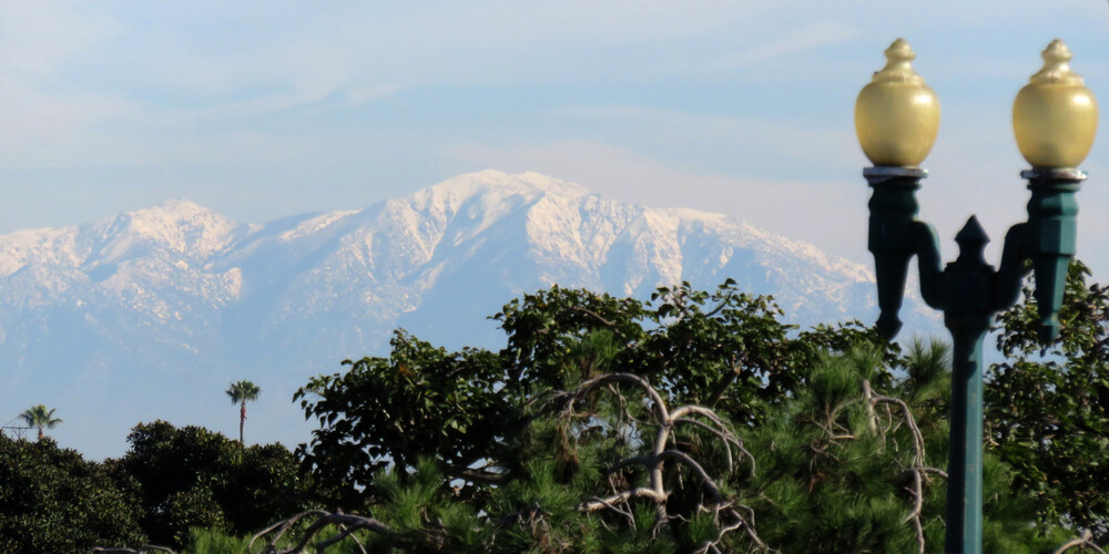 Snow-covered mountains in the distance, with partly cloudy sky above and green trees and a lamp post in the foreground.