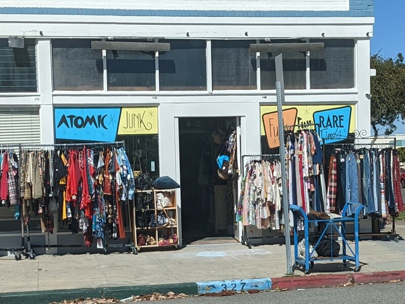 Thrift store with "Atomic Junk"  and "Rare Funky Finds" painted on its windows and racks of clothes out on the sidewalk.