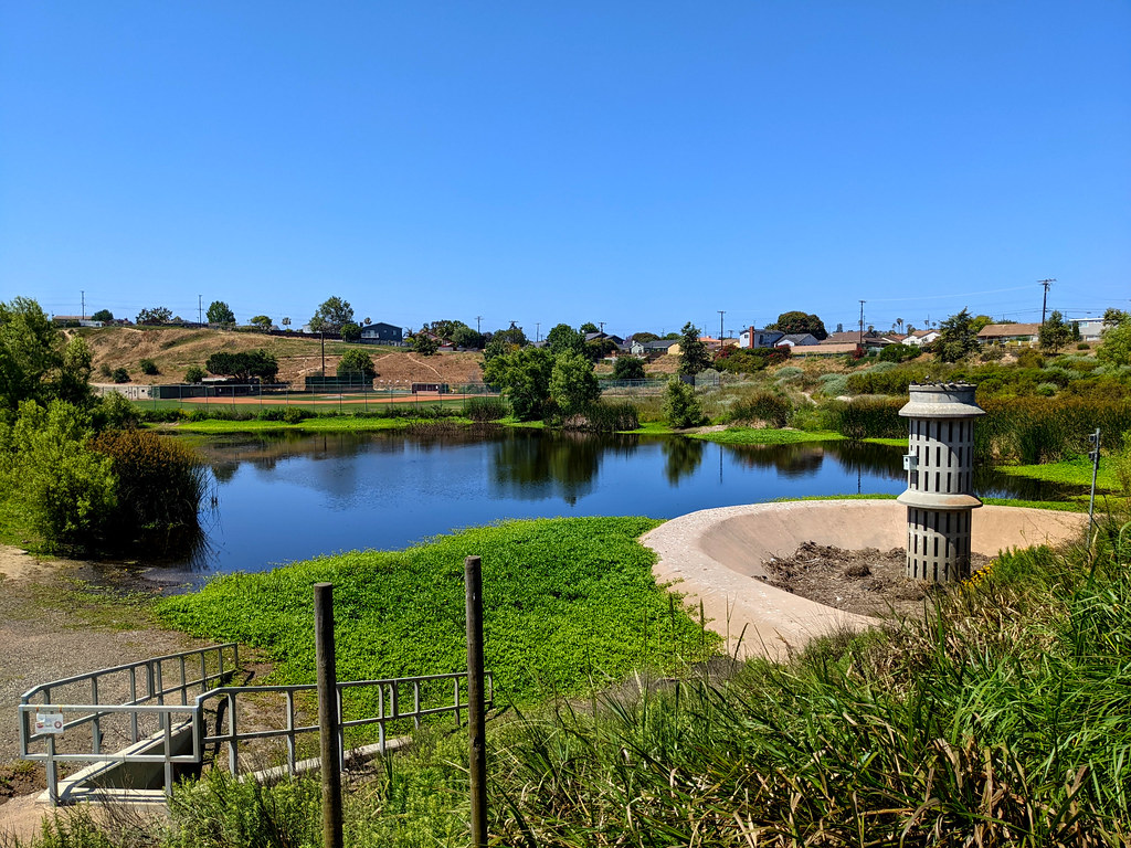 A smallish lake surrounded by green plants and golden brown hills, bright blue sky above it, and a large overflow drain in front of it off to the right.