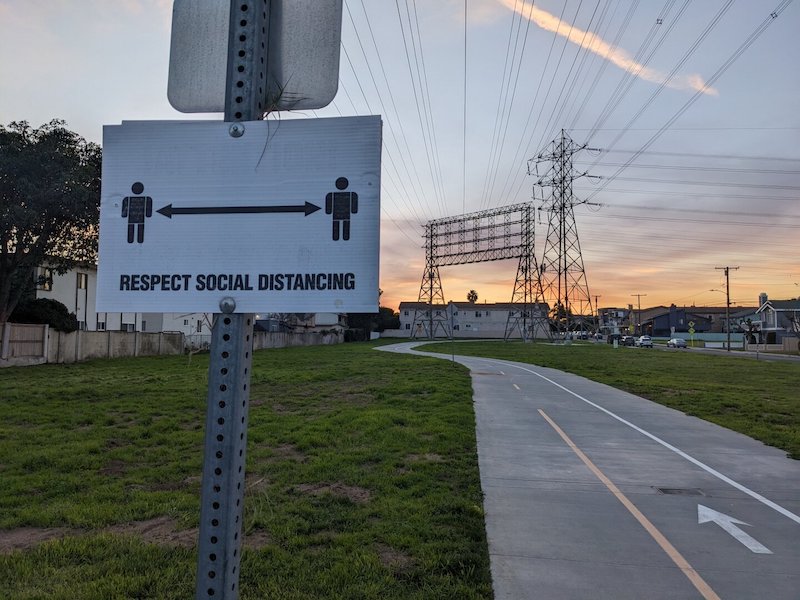 Bike path at dusk leading into the distance. up close is a sign with two stick figures and a double arrow between them, labeled RESPECT SOCIAL DISTANCING.