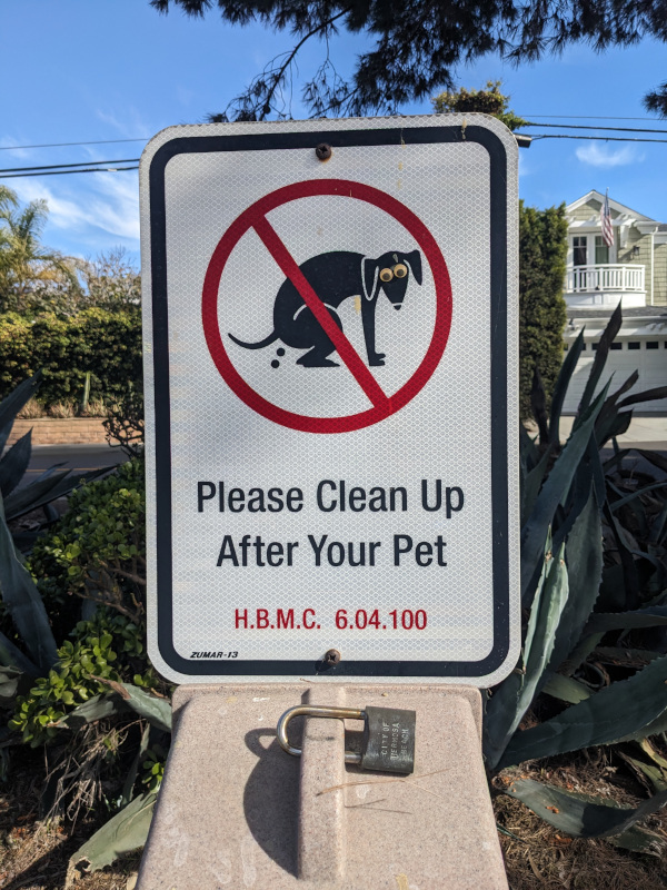 Sign saying Please Clean Up After Your Pet with a silhouette of a pooping dog and a red NO circle drawn around it. Someone has placed googly eyes on the dog.