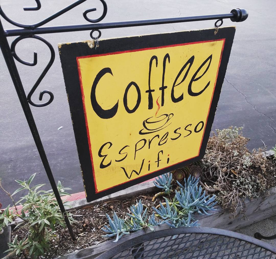 A squareish hand-painted metal sign with the words 'Coffee,' 'Espresso' and 'Wifi' stacked vertically. The letters and border are black on a bright yellow background. The sign hangs from an ornamented wrought-iron-style stand above a planter and is viewed diagonally from above. Between 'Coffee' and 'Espresso' there's a drawing of a cup and saucer with steam rising from it.