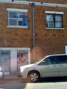 The same wall. The door's different, the paint on the Spider-Man figure is in better condition, and there's less graffiti around Superman, and there's a drainpipe next to the windows. You can see the reflection of someone holding up a phone because I didn't open the window, but otherwise not much has changed.