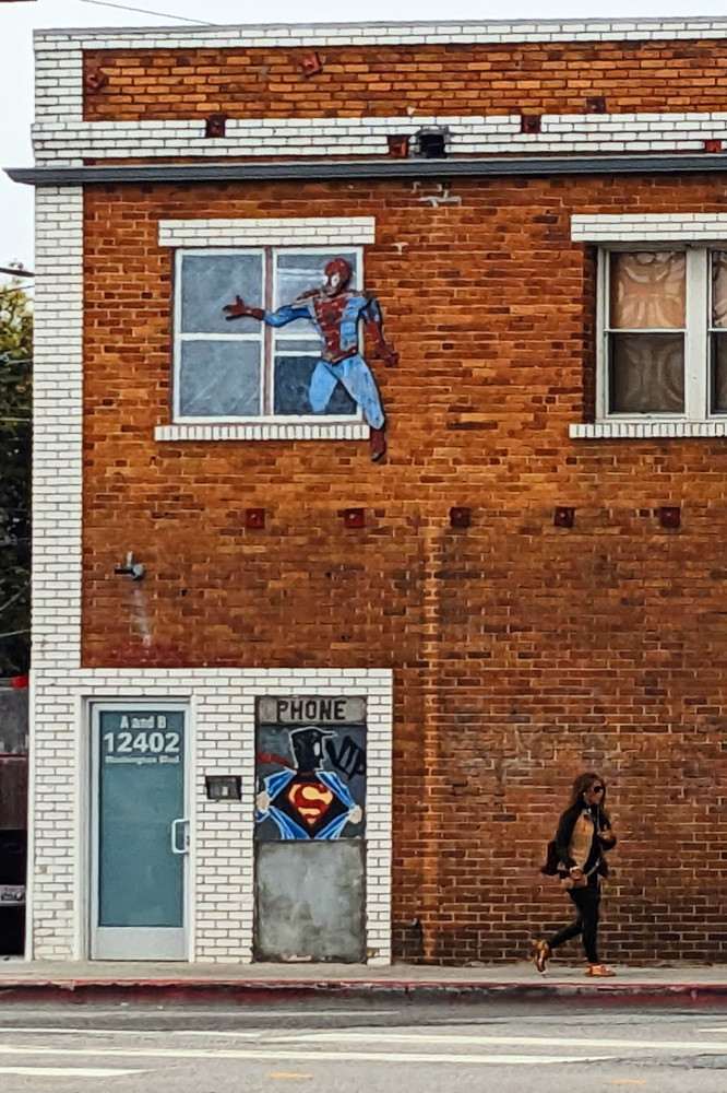 A red-brown brick wall. Two sets of windows are upstairs, and a door downstairs. Next to the door is a painted silhouette of a man pulling his outer shirt open and revealing the Superman symbol beneath it. The window above it has a scratched-up painted figure of  Spider-Man stepping out of the window. On the sidewalk below, a (real) woman walks by.