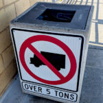 Square trash bin almost exactly the width of the road sign that's been attached to it, showing a silhouette of a large truck with a NO symbol over it, and the label OVER 5 TONS.