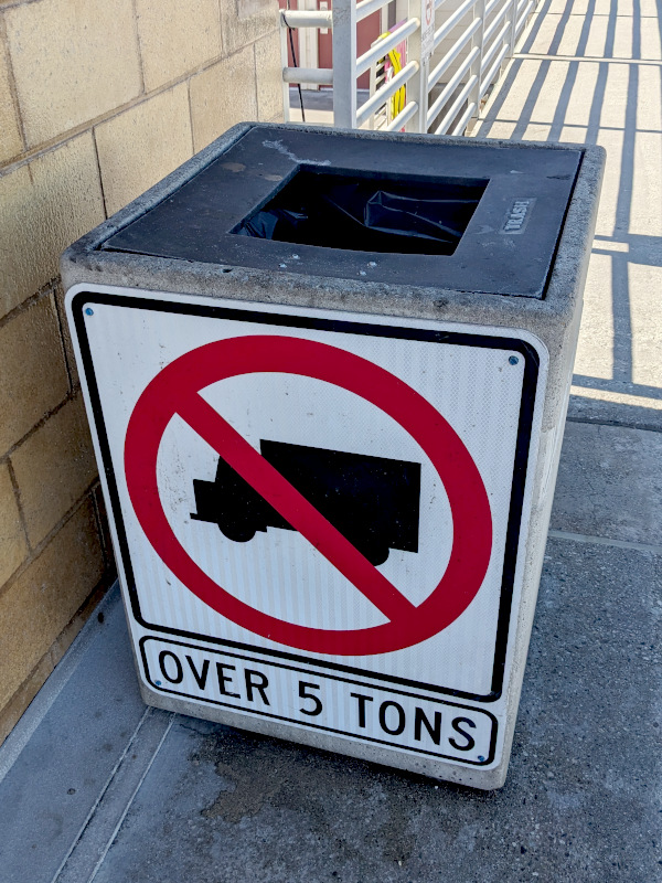 Square trash bin almost exactly the width of the road sign that's been attached to it, showing a silhouette of a large truck with a NO symbol over it, and the label OVER 5 TONS.