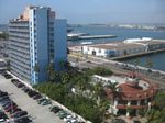 A somewhat tall, pale blue building with balconies lining one side and palm trees at the base, seen on a bright sunny day. A lower, round building is visible in the foreground. Beyond it are a street, some more parking, then docks with warehouses and a still harbor. The far shore is also visible, with lower buildings near the shore and low hills fading into the haze in the distance.