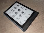 A slightly-bigger-than-hand sized flat black device with a light gray display showing a grid of application icons including Kindle, Kobo, KOReader, eBook Reader and more. A toolbar at the bottom of the display includes Library and Store along with Apps, Storage and Settings.