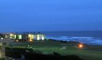 Looking down a green field to the coast at night. A path winds toward the ocean. Low, lit buildings are in the distance. A bright orange light shines out near the end of the path.