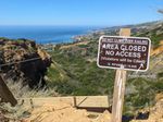 Looking past the edge of a cliff which runs off into the distance toward the ocean. There's a narrow wooden board at the edge of a trail, and a rope or cable running across the frame. In front of it, there's a post with a sign saying DO NOT CLIMB OVER RAILING. AREA CLOSED. NO ACCESS. (Violators will be Cited).