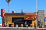 Small building with a tile roof, walls painted bright yellow. A red awning over the door proclaims EL TARASCO MEXICAN FOOD. Next to the door is a garage-sized roll-up glass door, with a patio area in front of it. There are no tables out - this was shortly before they opened. Cactus, succulents, and those weird orange fingery plant whose name I keep forgetting line the patio and the building. A sidewalk separates it from the street surface, where you can see room for one car to park between red-painted curbs, and someone has written HULK on the side of the curb. It's a bright sunny day, with a clear blue sky. One palm tree is visible behind the building, and an old, rounded-style car is parked around the corner. (There's a repair shop across the street.)