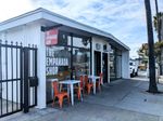 The front of a low building with an extremely shallow gabled roof, right off the sidewalk, with room for two storefronts. On the full-height windows in white, and hanging from a smaller, red sign facing the sidewalk, are the words THE EMPANADA SHOP. Two small white tables, each with a pair of orange chairs, sit in front of the window, and you can see several stools inside.