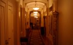 Looking along a hallway with a long geometric carpet over a wood floor, arches and columns spaced every so often, a few decorative columns against the walls, all dimly lit by yellowish, dish-shaped lamps hanging from the ceiling.