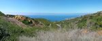 A panorama shot of mostly green hills, with some dry brush in the foreground and the ocean beyond. A valley curves out of view toward the ocean, what looks like a paved road running along the floor. (It's not - it's actually plastic ground cover. I'm not entirely sure what it's for, but it's a good bet it's related to mitigating the slow-motion landslide that's closed the area to the right due to unstable ground). Off to the left a steeper, bare section of hill is recognizable as the ridge from the first photo in this set.