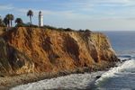 Red rugged cliffs rise above a pebbly beach while waves gently roll in from the ocean. A white column of a lighthouse sits atop the cliff, flanked by palm trees.